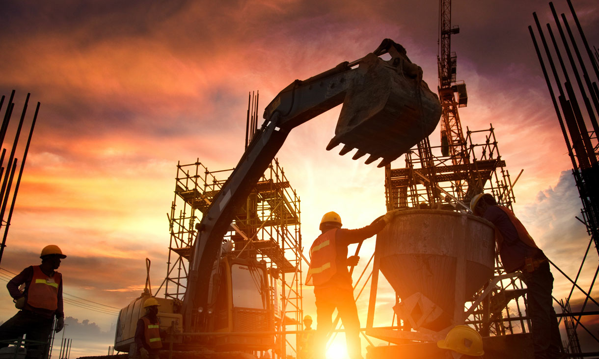 Construction workers operating machinery while the sun sets over the site.